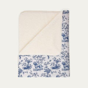 Peut inclure: Un plaid plié présentant un motif toile de Jouy bleu et blanc avec des illustrations de chiens et d'arbres. La couverture possède une doublure en polaire sherpa couleur crème, visible sur la partie repliée, contrastant avec l'extérieur imprimé.