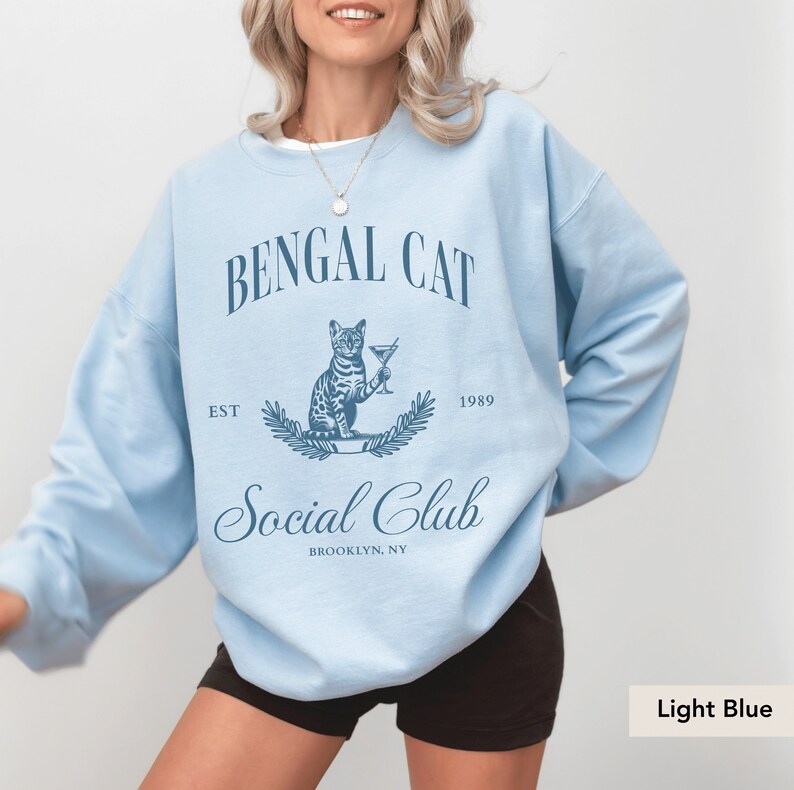 May include: Light blue sweatshirt with the text "BENGAL CAT Social Club" and a graphic of a Bengal cat holding a cocktail. The text "EST 1989" and "BROOKLYN, NY" are also printed on the sweatshirt. The sweatshirt is a solid light blue color.