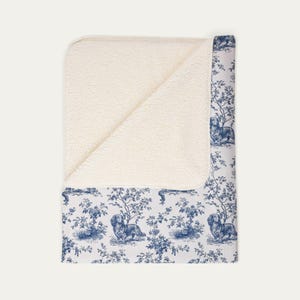 May include: A rectangular throw blanket featuring a white, textured, fleece-like underside and a top side with a blue and white toile pattern. The pattern displays repeating illustrations of dachshund dogs positioned among detailed trees and floral foliage.