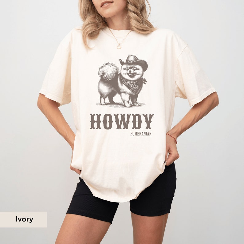 May include: Ivory t-shirt featuring a graphic of a Pomeranian dog wearing a cowboy hat and bandana. The text "HOWDY POMERANIAN" is printed below the dog.