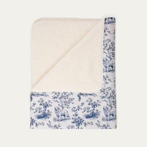 Peut inclure: Une couverture jeté pliée présentant un intérieur en polaire sherpa couleur crème et un extérieur blanc avec un motif toile de Jouy bleu répétitif représentant des chiens parmi des arbres et du feuillage. La couverture est partiellement pliée.