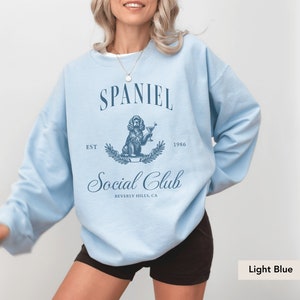 May include: Light blue sweatshirt with a graphic of a spaniel dog holding a martini glass. The text "Spaniel Social Club" is printed on the sweatshirt, along with "Est. 1986" and "Beverly Hills, CA".