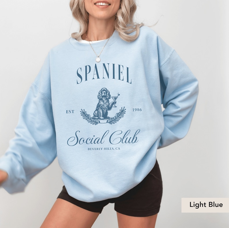 May include: Light blue sweatshirt with a graphic of a spaniel dog holding a martini glass. The text "Spaniel Social Club" is printed on the sweatshirt, along with "Est. 1986" and "Beverly Hills, CA".