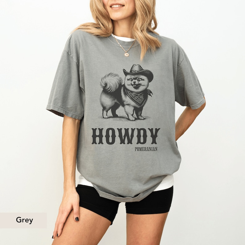 May include: A grey t-shirt featuring a black and white illustration of a Pomeranian dog wearing a cowboy hat and bandana. The text "HOWDY POMERANIAN" is printed below the dog.