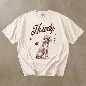 Puede incluir: Camiseta color crema con una ilustración marrón de un perro con sombrero de vaquero y pañuelo. La palabra "Howdy" está escrita encima del perro en una fuente estilizada, con estrellas.