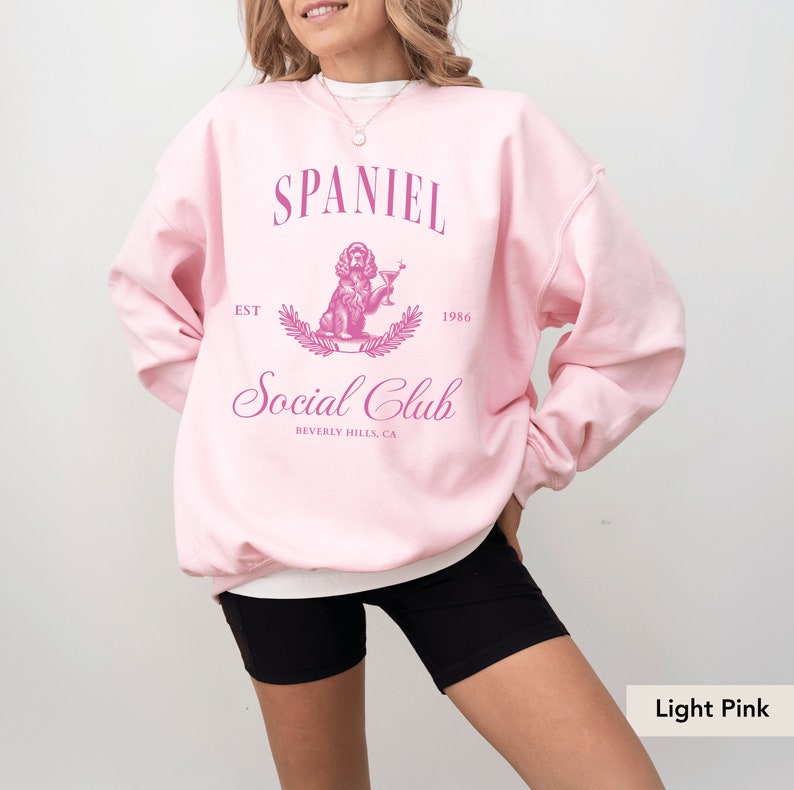May include: Light pink sweatshirt with a pink graphic of a spaniel dog holding a martini glass. The graphic says "Spaniel Social Club" with the text "Est. 1986" and "Beverly Hills, CA".