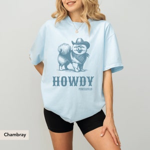 May include: A light blue t-shirt featuring a graphic of a Pomeranian dog wearing a cowboy hat and bandana. The text "HOWDY" and "POMERANIAN" is printed below the dog. The shirt is called " Chambray".