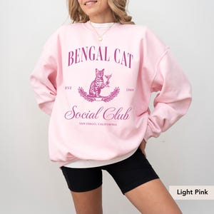 May include: Light pink sweatshirt with the text "BENGAL CAT Social Club" and a graphic of a Bengal cat holding a cocktail. The text includes "EST 1989" and "SAN DIEGO, CALIFORNIA".