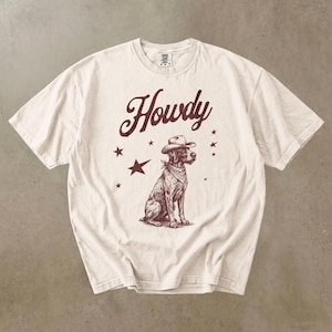 Puede incluir: Camiseta de color crema con una ilustración marrón de un perro con sombrero de vaquero y pañuelo. La palabra "Howdy" está escrita encima del perro en una fuente estilizada, con estrellas marrones dispersas alrededor del diseño.