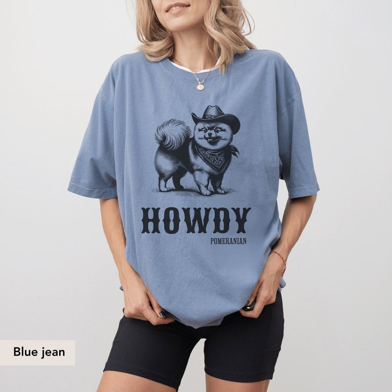 May include: A blue jean t-shirt featuring a black and white illustration of a Pomeranian dog wearing a cowboy hat and bandana. The text "HOWDY" and "POMERANIAN" is printed below the dog.