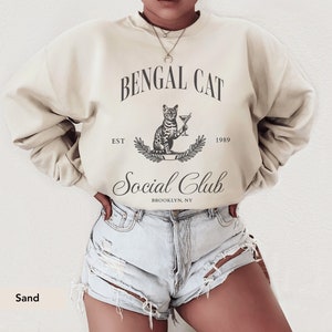 May include: Sand-colored sweatshirt with a graphic that reads "Bengal Cat Social Club" with a cat illustration holding a martini. The text "EST 1989" and "Brooklyn, NY" are also included in the design. The sweatshirt is a neutral color.