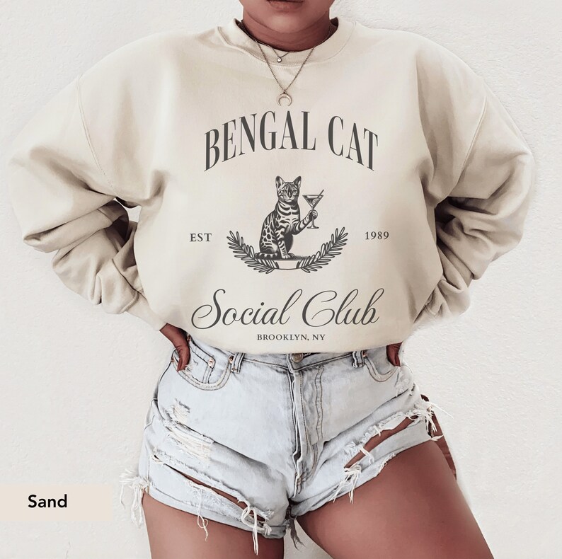 May include: Sand-colored sweatshirt with a graphic that reads "Bengal Cat Social Club" with a cat illustration holding a martini. The text "EST 1989" and "Brooklyn, NY" are also included in the design. The sweatshirt is a neutral color.