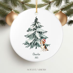 May include: A white ceramic Christmas ornament featuring a watercolour illustration of a snow-covered fir tree and a beagle wearing a Santa hat. The name "Charlie" and the year "2025" are printed below the dog.