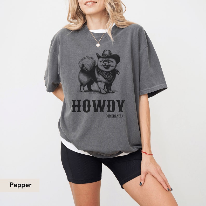 May include: A grey t-shirt featuring a black and white illustration of a Pomeranian dog wearing a cowboy hat and bandana. The text "HOWDY POMERANIAN" is printed below the dog. The model is wearing black cycling shorts.