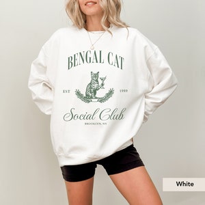 May include: White sweatshirt with a green graphic that reads "Bengal Cat Social Club, Brooklyn, NY." The graphic features a cat holding a cocktail glass. The sweatshirt has a crew neck and long sleeves.