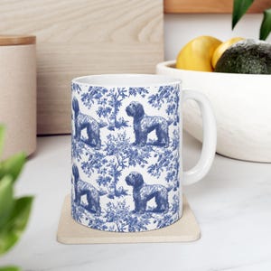 May include: White ceramic mug with a blue and white pattern of dogs and floral designs. The mug has a handle and is sitting on a beige coaster.