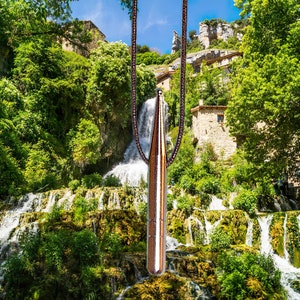 May include: A copper-colored pendant necklace with a bullet-shaped charm hangs in front of a cascading waterfall. The charm is elongated and metallic, suspended from a brown chain. The background features lush green foliage, stone buildings, and a bright blue sky.
