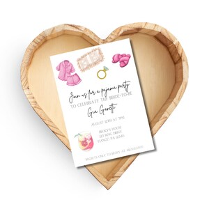 May include: A white invitation card with watercolor illustrations of pajamas, slippers, and a ring, inside a heart-shaped wooden tray. The invitation text reads "Join us for a pajama party to celebrate the bride-to-be, Gia Garetti."