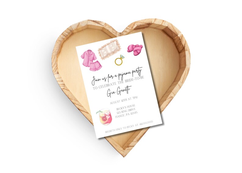 May include: A white invitation card with watercolor illustrations of pajamas, slippers, and a ring, inside a heart-shaped wooden tray. The invitation text reads "Join us for a pajama party to celebrate the bride-to-be, Gia Garetti."