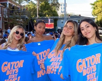 May include: Four people holding up blue t-shirts with the text "GATOR BOYS STAY HOT!" in orange. The image is taken outdoors on a sunny day, with a crowd and buildings in the background.