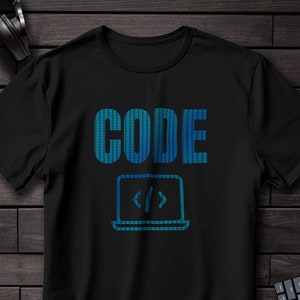 Puede incluir: Camiseta negra con la palabra "CODE" en fuente pixelada azul sobre un icono de portátil con corchetes de código. La camiseta está sobre una superficie de madera oscura, con auriculares, un cuaderno, gafas y un teclado.