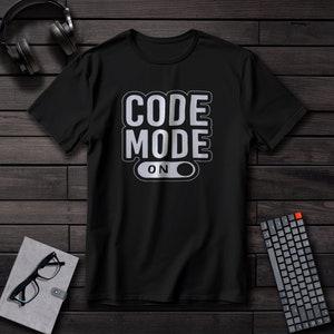 Puede incluir: Camiseta negra con el texto "CODE MODE ON" en diseño blanco y gris. La camiseta se muestra sobre una superficie de madera oscura, con auriculares, un teclado, gafas y un cuaderno en el fondo.