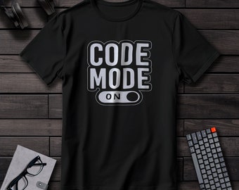 May include: Black t-shirt with the text "CODE MODE ON" in a white and gray design. The shirt is displayed on a dark wood surface, with headphones, a keyboard, glasses, and a notebook in the background.