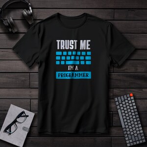 Puede incluir: Camiseta negra con el texto "TRUST ME I'M A PROGRAMMER" en blanco y azul. El diseño incluye un gráfico de teclado. La camiseta se muestra sobre una superficie de madera oscura, con auriculares, un teclado y un cuaderno con gafas.