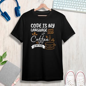 Könnte beinhalten: Schwarzes T-Shirt mit dem Text "CODE IS MY LANGUAGE and Coffee IS MY FUEL". Das Design umfasst eine Kaffeetasse und Kaffeebohnen-Illustrationen. Das T-Shirt hängt an einem Holzbügel.