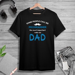 Puede incluir: Camiseta negra con el texto "SOME PEOPLE CALL ME PROGRAMMER the most important calls me DAD" en blanco y azul. La camiseta está colgada de una percha de madera.