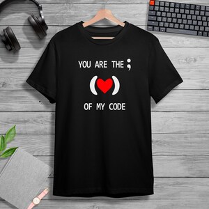 Puede incluir: Camiseta negra con el texto blanco "YOU ARE THE ; OF MY CODE". El diseño incluye un símbolo de corazón rojo entre paréntesis. La camiseta está colgada de una percha de madera.