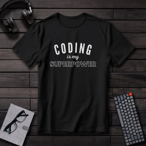 Puede incluir: Camiseta negra con el texto blanco "CODING is my SUPERPOWER". La camiseta se muestra sobre una superficie de madera oscura, junto con un teclado, auriculares y un cuaderno con gafas.
