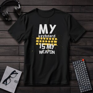 Puede incluir: Camiseta negra con el texto "MY keyboard IS MY WEAPON" en blanco y amarillo. El diseño incluye un gráfico de teclado amarillo. También se ven unas gafas, un cuaderno y un teclado.