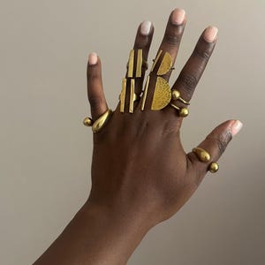 以下が含まれることがあります： 金色の指輪のコレクションを展示する手。リングは、バー、半円、球など、さまざまな幾何学模様を特徴としています。リングは複数の指に着用され、さまざまな形とサイズを示しています。画像には「MZURI STUDIO」というテキストが含まれています。