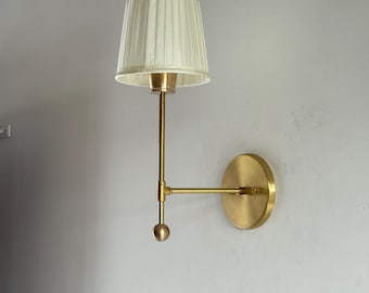 Könnte beinhalten: Wandleuchte aus Messing mit cremefarbenem, plissiertem Lampenschirm. Die Leuchte hat einen runden Sockel und einen schlanken Arm mit einem kleinen Kugeldetail. Geeignet für die Innenbeleuchtung.