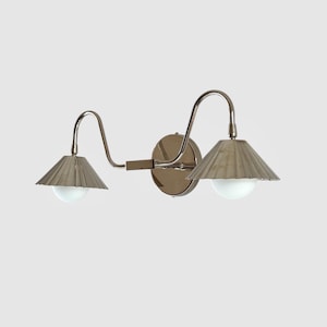 Op de afbeelding: Een wandlamp met twee lampen met een metallic afwerking. De lamp heeft twee gebogen armen, elk met een conische kap met een getextureerd oppervlak. Elke kap heeft een witte, ronde gloeilamp. De lamp is op een cirkelvormige basis gemonteerd.