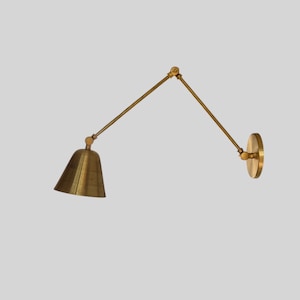 Op de afbeelding: Een messing wandlamp met een verstelbare arm en een conische kap. De lamp heeft een ronde wandplaat en een draaibare arm voor gerichte verlichting. De afwerking is een warm, metallic goud.