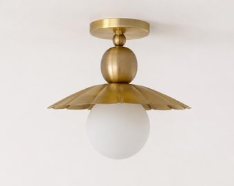 May include: A flush mount ceiling light fixture featuring a circular brass canopy, a central spherical brass accent, and a wide, scalloped brass shade. A single, spherical frosted white glass globe bulb is suspended beneath the metal shade.