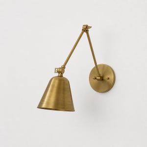 May include: A wall-mounted lamp with a brass finish, featuring a circular wall plate, an adjustable articulated arm with two segments, and a downward-facing conical shade. The metal construction has a brushed texture throughout the entire fixture.