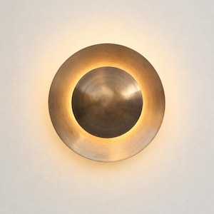 Pode incluir: Um candeeiro de parede circular com acabamento em bronze escovado e um elemento central em forma de cúpula. O design incorpora uma fonte de luz integrada que cria um brilho suave e quente que emana de trás da cúpula central, iluminando o anel exterior.