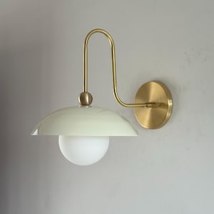 May include: A modern wall sconce with a pale green dome shade and a frosted globe light. The fixture has a curved, brass-colored arm and a round mounting plate. The design is minimalist and elegant, suitable for various interior styles.