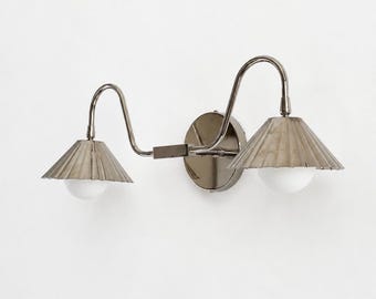 May include: A double wall sconce featuring a polished nickel finish with two curved arms extending from a central circular mounting plate. Each arm supports a pleated metallic cone shade and a round white glass globe bulb for illumination.