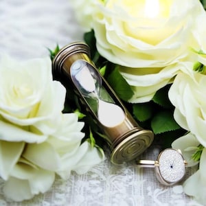 May include: A brass hourglass with white sand sits on a white lace surface surrounded by white roses.