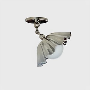 May include: A contemporary ceiling light in polished chrome. It has a circular base, a curved arm with ball joints, and a shade designed to look like a ruffled, metallic fabric. A white, spherical light bulb is visible within the shade.