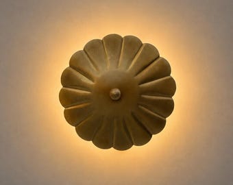 May include: A circular, brass-toned wall light fixture with a scalloped, petal-like design radiating from a central knob. The fixture emits a soft, warm glow from behind its edges, creating a halo effect against the surrounding surface.