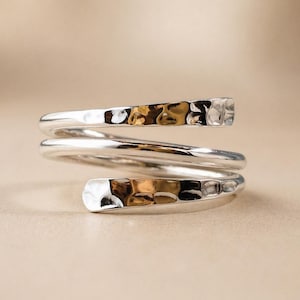 Puede incluir: Un anillo envolvente plateado con textura martillada. El anillo se enrolla alrededor del dedo, con extremos planos y rectangulares. La textura martillada refleja la luz, creando reflejos.