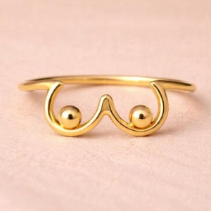 Peut inclure: Une bague dorée au design unique. La bague présente deux sphères circulaires et une courbe centrale pointant vers le haut, créant une forme stylisée. La bague est posée sur un fond rose clair.