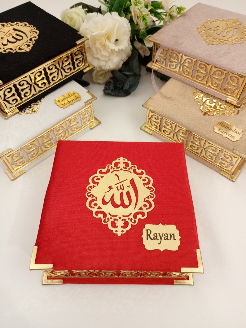 May include: Three decorative boxes with gold trim and intricate designs. The boxes are covered in velvet fabric in black, pink, and red. The boxes have a gold inscription of the Arabic word for God, "Allah", and a gold label with the text "Ramadan Mubarak", "Ramadan Kareem", and "Rayan".