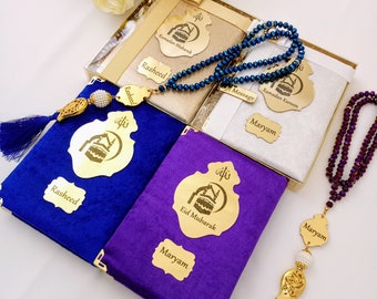 Könnte beinhalten: Ein Set aus zwei Gebetsbüchern mit goldenen Akzenten und Quasten, eines blau und eines lila, mit den Namen "Rasheed" und "Maryam" darauf. Die Bücher befinden sich in einer Geschenkbox mit einem goldenen Band. Es gibt auch zwei Gebetsperlen, eine blaue und eine lila, mit goldenen Akzenten und Quasten. Die Perlen tragen die Namen "Rasheed" und "Maryam".