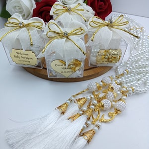Op de afbeelding: Witte geschenkdozen met gouden lint en een transparant venster, gevuld met witte parels en een gouden hartbedel. De dozen hebben gepersonaliseerde etiketten met de tekst "Welcome Baby Suriayah 12.12.2023", "Omar & Sarah's Nikkah 12.12.2023" en "Yousouf & Sarah's Nikkah 12.12.2023". Een witte parelketting met gouden accenten en kwastjes.